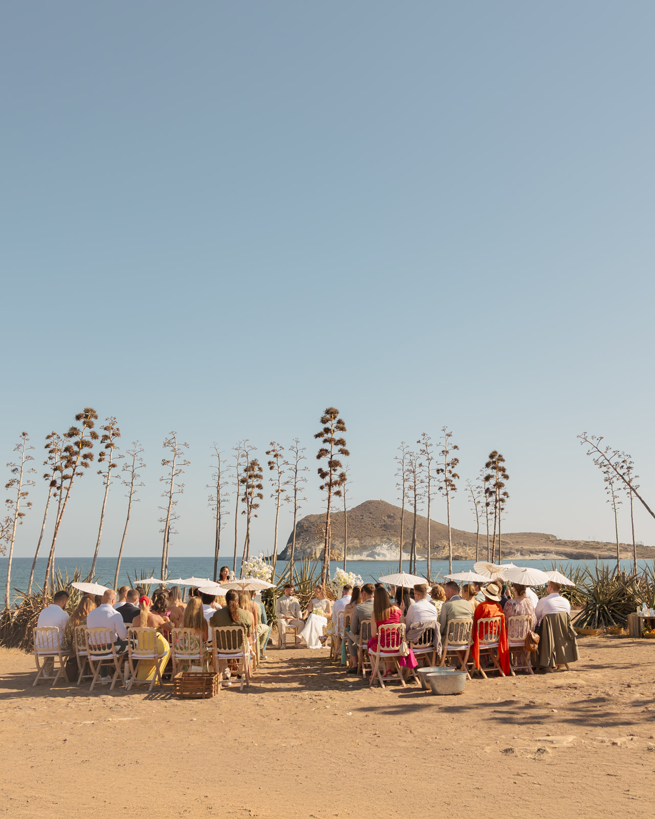 Freie Trauung am Strand von San Jose - Gaeste mit Papier-Sonnenschirmen, hohe Agaven, Meer und Felsen von Cabo de Gata