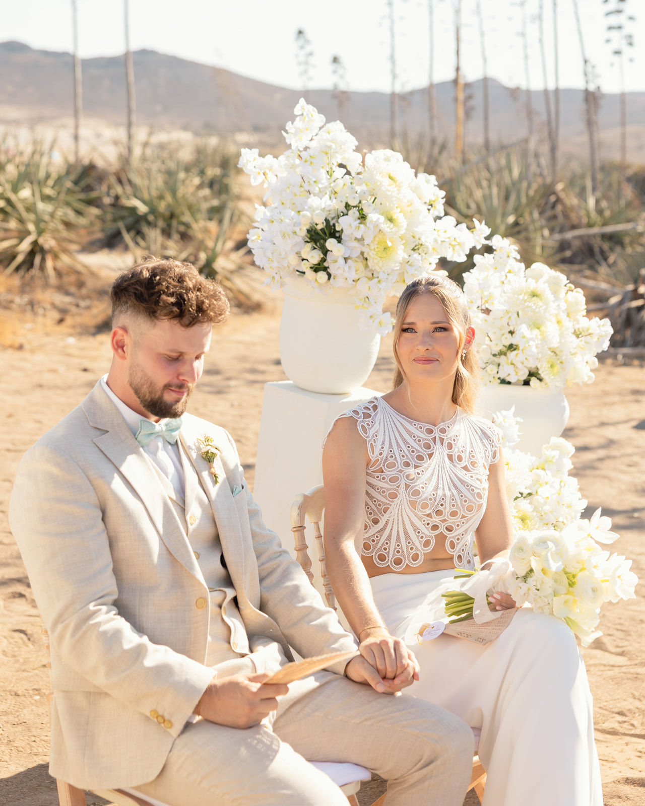 Katharina und Roman sitzen waehrend der Zeremonie auf dem Sand, umgeben von weissen Blumenarrangements, Wuestenlandschaft Cabo de Gata