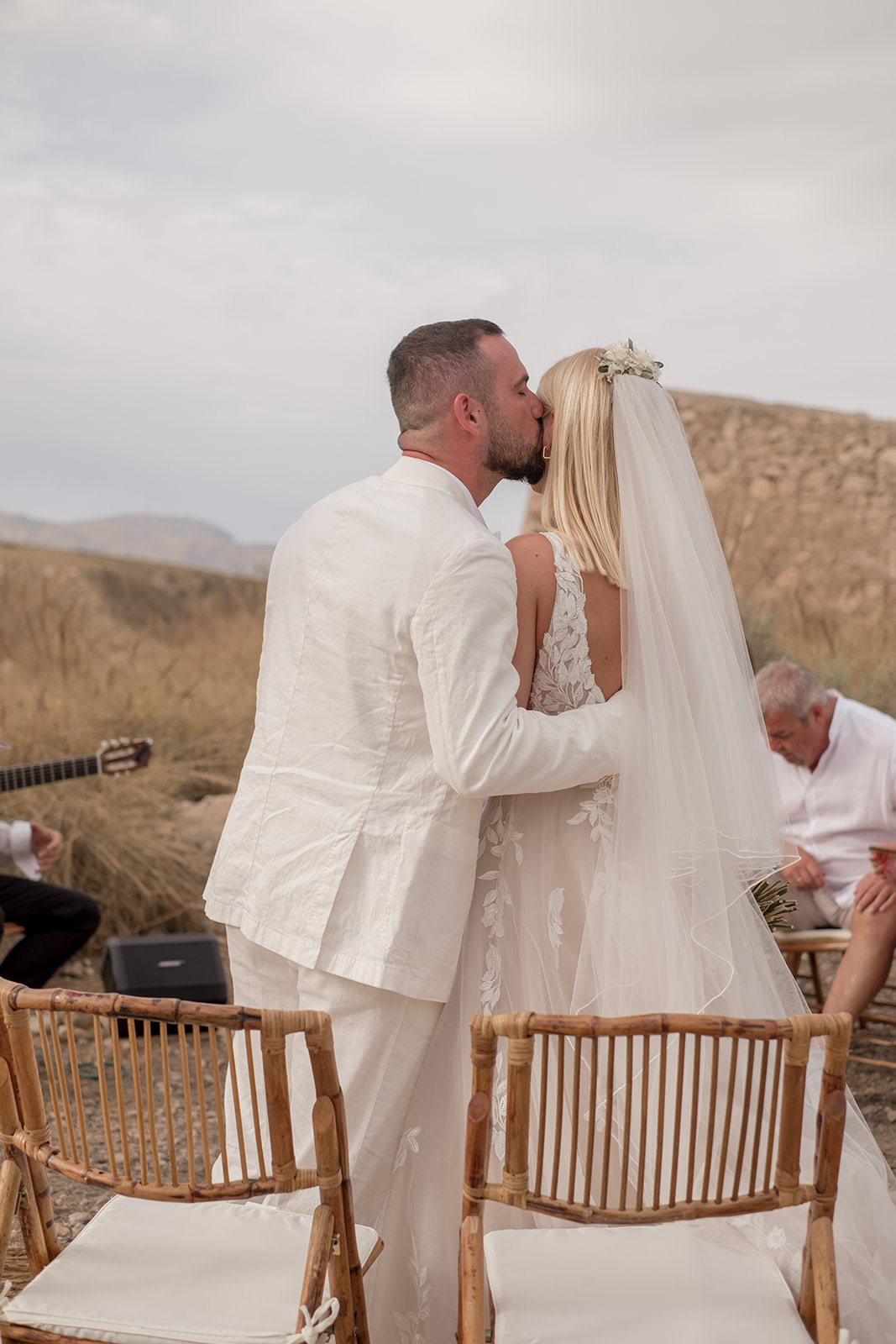 Lara und Michael küssen sich von hinten fotografiert, Rattanstühle der Gäste und Gitarrist im Hintergrund