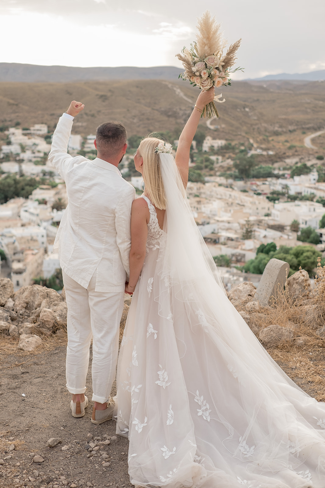 Lara und Michael von hinten auf dem Felsgipfel über Agua Amarga, beide recken die Fäuste in die Luft – Lara hält ihren Pampasgras-Bouquet hoch