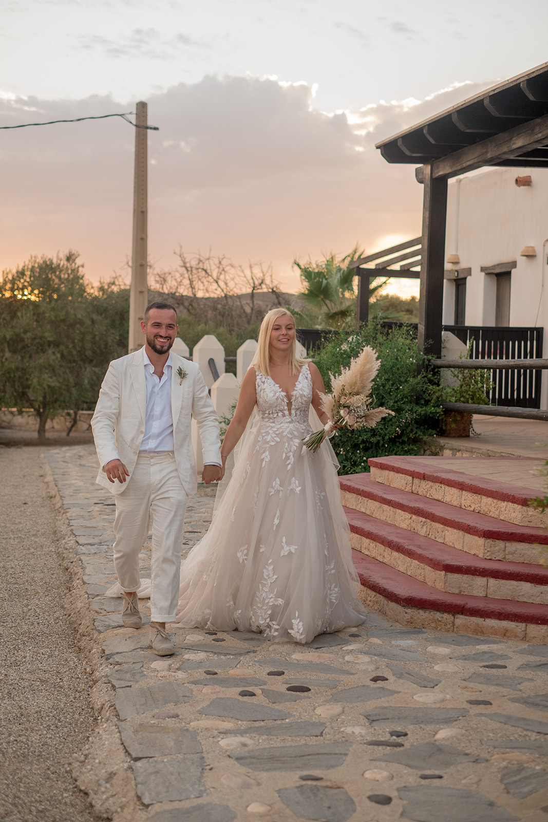 Lara und Michael gehen Hand in Hand bei Sonnenuntergang über einen Steinpfad der andalusischen Finca