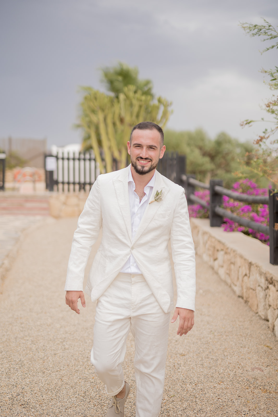 Bräutigam Michael im cremefarbenen Leinenanzug mit Eukalyptus-Boutonnière, lächelnd auf dem Kiesweg der Finca, Bougainvillea im Hintergrund
