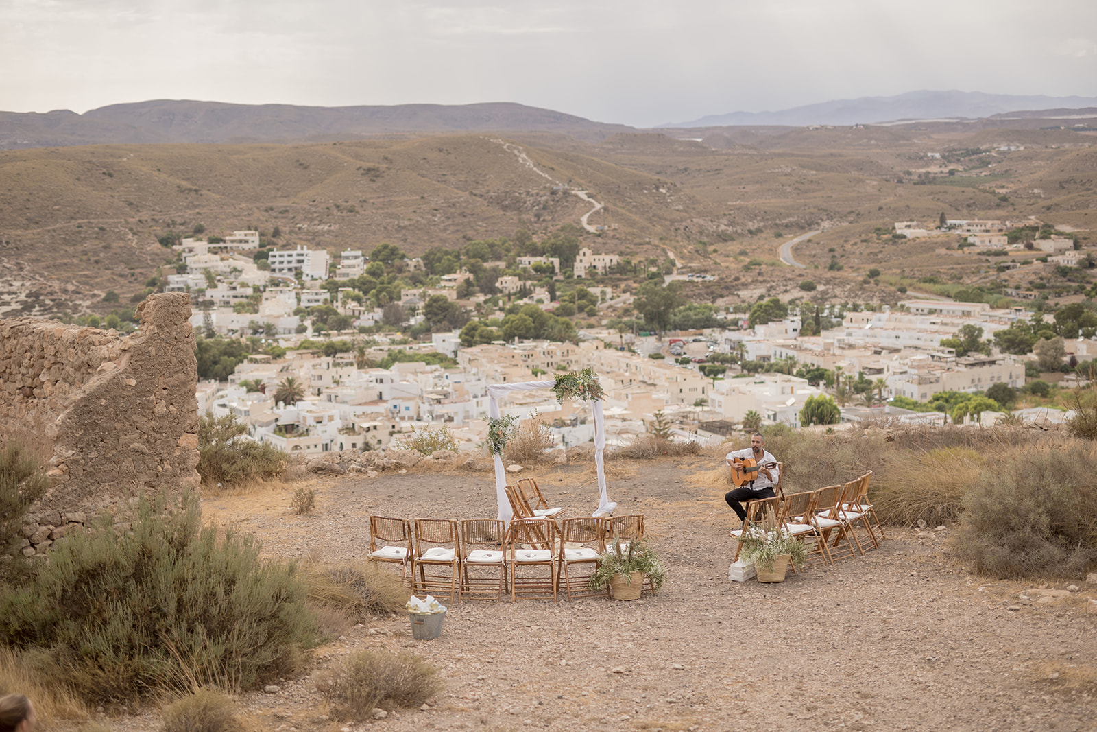 Hochzeits-Setup auf dem Felshügel über Agua Amarga: Rattanstühle im Halbkreis, weißer Traubogen, Gitarrist – Panoramablick auf Dorf und Mittelmeer