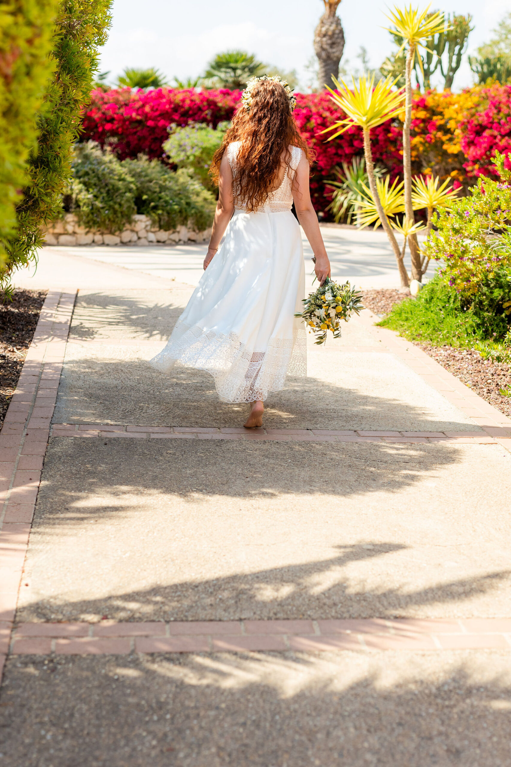 Braut mit rotem Lockenhaar und Blumenkranz im Boho-Spitzenkleid geht durch den Garten, Agua Amarga, Boho Strandhochzeit Cabo de Gata