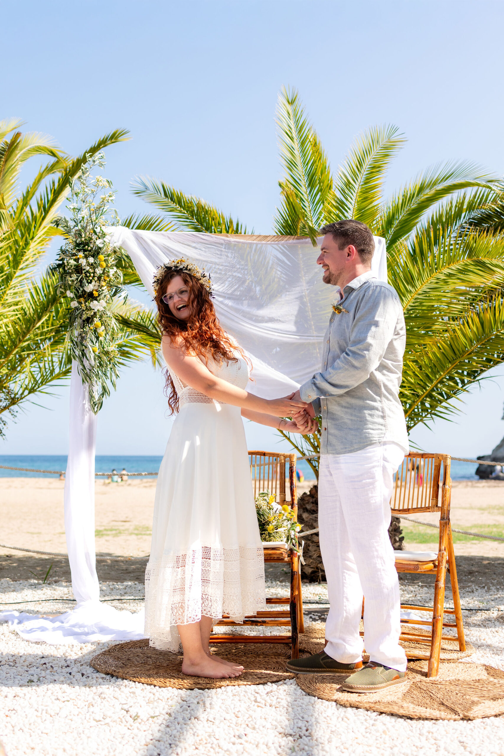 Brautpaar hält lachend Händchen unter dem Blumenbaldachin am Strand, Cabo de Gata, Boho Strandhochzeit